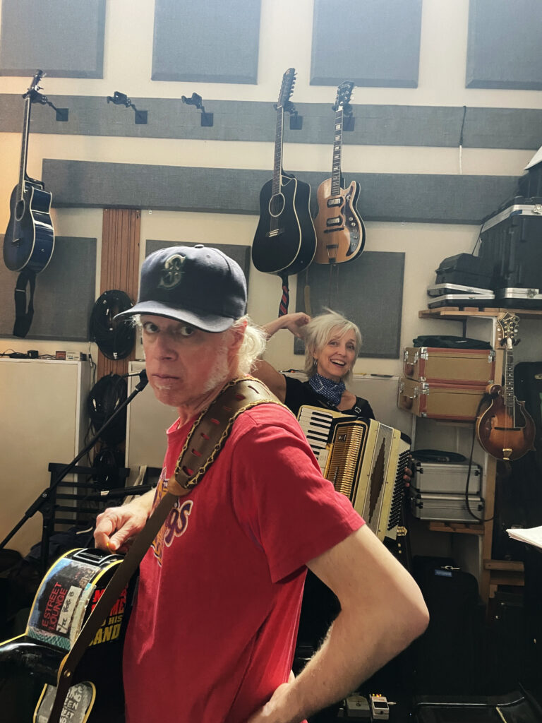 Foster Motel in their rehersal space surrounbded by instruments and cases; Joel Stewart in foreground holding his black acoustic guitar, with Jane Gowan in the background holding an accordion